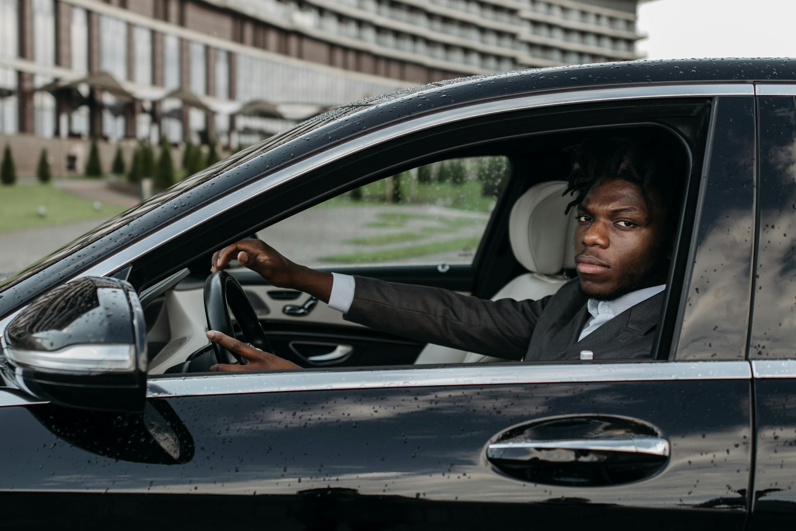 African American man confidently driving a sleek black luxury car outside a modern building.