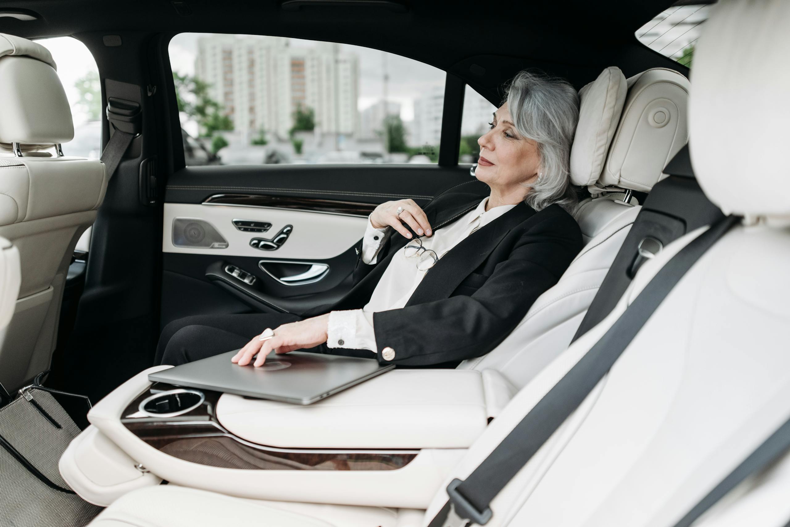Senior woman in a luxury car with a laptop, gazing out the window, representing business travel.